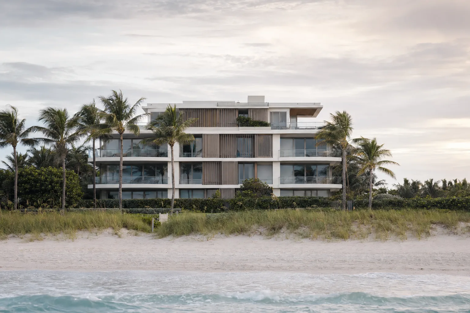 A low-rise oceanfront residential building in Surfside seen from the beach at early morning, calm Atlantic surf, manicured dune grass, soft overcast light, no people in frame.